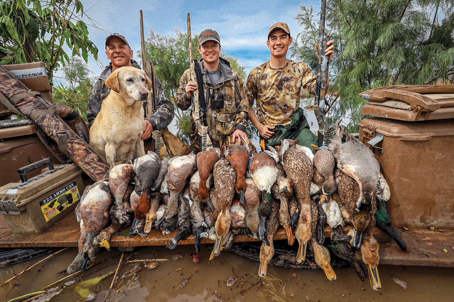 three hunters and yellow lab pose behind large pile of dead waterfowl