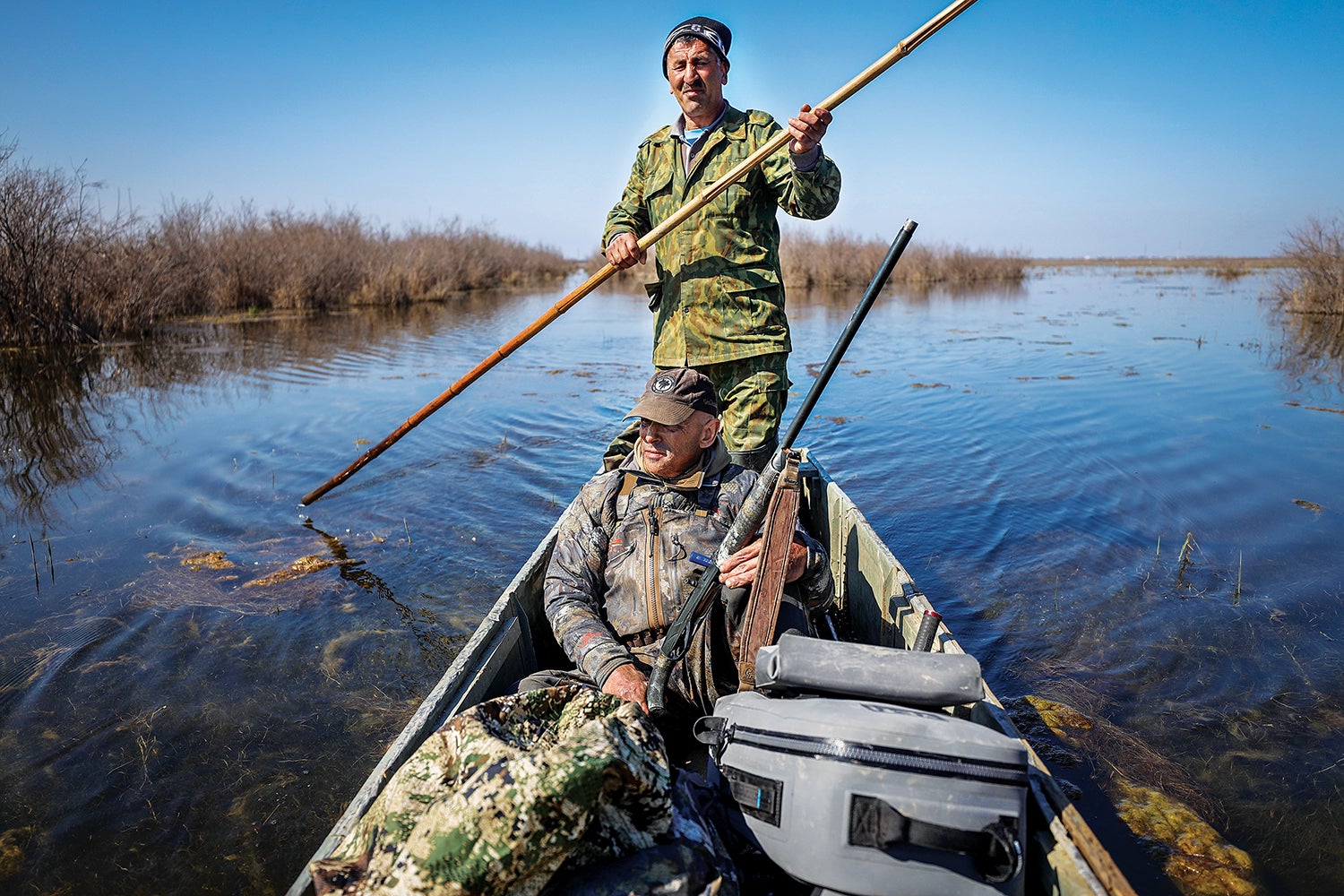 duck hunter sits in boat being push-poled by azerbaijani man through swampy area